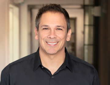 Jamey Schrier, a business coach for private practice owners, smiling in a professional headshot. He is wearing a black collared shirt, with short, dark hair, and a warm expression. The background is softly blurred, featuring an indoor hallway with natural lighting.